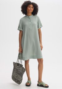 Hellgrünes, geripptes Kleid mit Stehkragen, kurzen umgeschlagenen Ärmeln und Knopfverschlüssen. Kombiniert mit einer geflochtenen Tasche und grünen Sandalen.