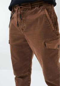 Bruna cargobyxor med elastisk midja, snörning och sidofickor. Slät textur med en subtil glans och skräddarsydd passform.