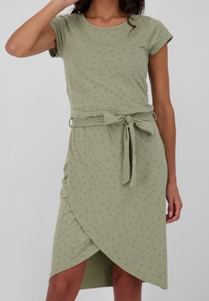 Kobieta w jasnzozielonej sukience z krótkim rękawem z małym ciemnym wzorem, przewiązanym paskiem w talii i asymetryczną spódnicą w stylu kopertowym.