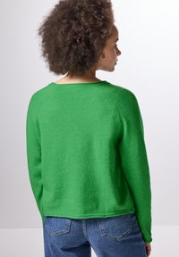 Grön stickad tröja med avslappnad passform, raglanärmar och bred halsringning, kombinerad med blå jeans. Subtil textur och fållavslut.