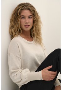 Pale bézs kötött pulóver kerek nyakkivágással, bordás ujjakból és laza fazonban. Fekete nadrággal párosítva, semleges háttér előtt.