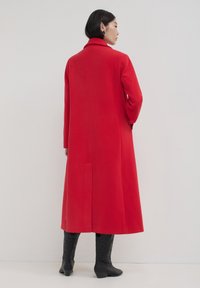 Femme de dos, portant un long manteau rouge et des bottes noires mi-mollet, sur un fond clair et uni.
