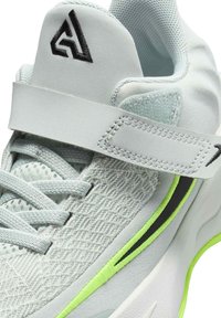 Zapatilla deportiva de color gris claro con parte superior de malla, acentos negros, una correa de Velcro y detalles en verde lima a lo largo de la suela. Tela texturizada.