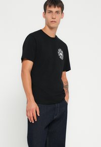 Svart bomull t-skjorte med korte ermer, med en sirkulær hvit grafikk på venstre bryst. Kombinert med mørk denimjeans.