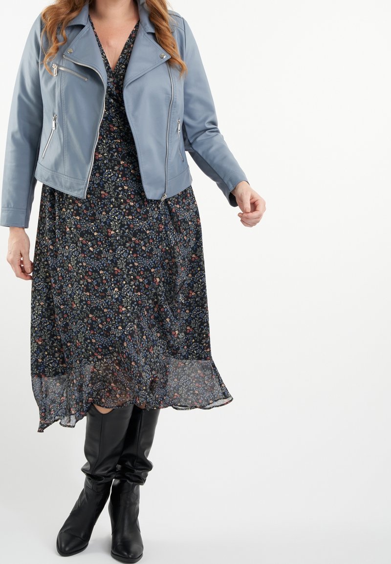 Veste de moto en cuir bleu portée sur une robe à fleurs foncées. La robe possède un ourlet fluide, et une paire de bottes noires montantes complète la tenue.