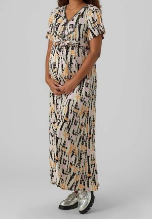 Gravid kvinde iført en mønstret maxikjole med korte ærmer og sølvfarvede dekorerede loafers, stående mod en grå baggrund.