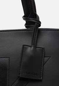 Sac fourre-tout en cuir noir avec texture lisse, comprenant une étiquette de logo, une fermeture éclair et des coutures ton sur ton. Agrémenté d'un design graphique.