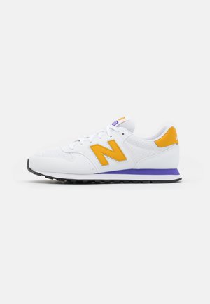 Biały sportowy sneaker z żółtym logo ""N"", żółtą naszywką na pięcie, fioletowym paskiem na podeszwie środkowej i czarną gumową podeszwą zewnętrzną.