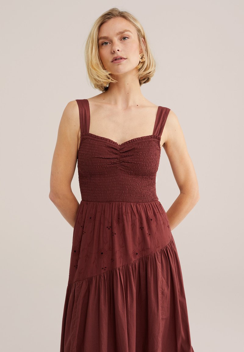Vrouw met kort blond haar, gekleed in een mouwloze bordeauxrode jurk met gesmokte lijf en bloemrijke borduursels op de rok, staand tegen een effen achtergrond.