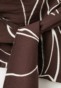Tissu marron avec des lignes courbes blanches abstraites nouées en un nœud lâche, montrant les détails de la texture et du motif.