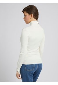 Maglione bianco a collo alto in maglia con design aderente, maniche lunghe e polsini a costine. Abbinato a jeans in denim blu, visto da dietro.