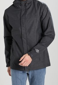 Giacca impermeabile nera con zip frontale, cappuccio regolabile e logo sul petto. Presenta due tasche laterali e una trama sottile.