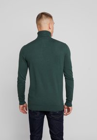 Dunkelgrüner langärmeliger Rollkragenpullover mit geripptem Hals und Bündchen, strukturierter Stoff und schlichtem Design. Getragen mit dunklen Jeans.