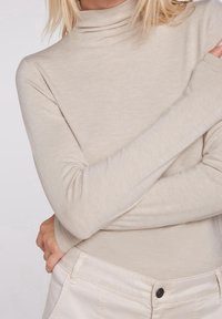 Beige kauluspaita, joka on valmistettu pehmeästä, kevyestä kankaasta ja jossa on pitkät hihat. Malli on poseerannut käsivarret ristissä, esitellen tyylikästä vartalonmyötäistä leikkausta.