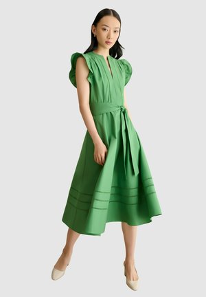 Mujer con vestido midi verde sin mangas, con volantes en los hombros y cinturón en la cintura, combinado con tacones beige de punta fina.