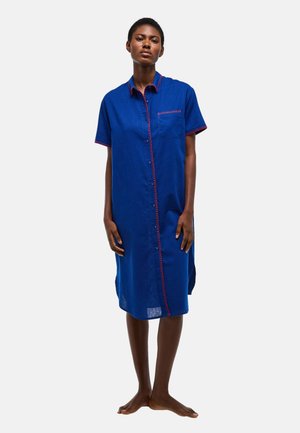 Femme debout pieds nus portant une robe bleue boutonnée au genou avec des coutures rouges et une poche poitrine, sur un fond clair uni.