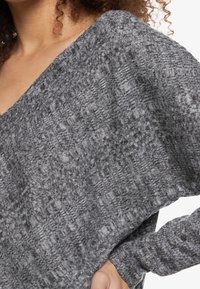 Suéter gris texturizado con un escote ancho y mangas largas. La tela presenta un patrón entrelazado y una apariencia suave y ligeramente pelusa.