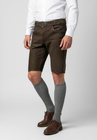 Braune Leder-Shorts mit dekorativer Stickerei, silbernen Beschlägen und einem Knopfdetail, kombiniert mit grauen knielangen Socken und braunen Schuhen.