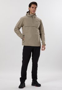 Veste imperméable beige avec capuche, poche avant et cordons de serrage réglables ; assortie à un pantalon noir et des chaussures noires. Texture lisse.