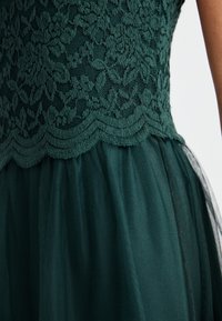 Grön spetsblus med scalloped kanter och blommönster, kombinerad med en lager av tyllkjol i en mörkare grön nyans. Delikat textur totalt.