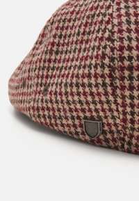 Barna és piros houndstooth mintázatú gyapjú sapka, sötét fém logóval ellátott folttal, kerek formával és enyhén texturált felülettel.