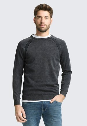 Mann mit kurzen Haaren und Bart, trägt einen dunkelgrauen Pullover über einem weißen Hemd und blauen Jeans, steht mit einer Hand in der Tasche.
