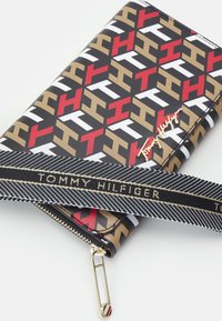 Czarny portfel z geometrycznym wzorem w czerwonym, białym i złotym kolorze, z logo w złotym kolorze oraz czarnym paskiem w paski z napisem "TOMMY HILFIGER."