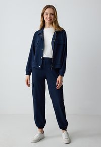 Marineblauwe trainingspak gemaakt van zachte stof. Het jack heeft een ritssluiting en twee borstzakken; de broek heeft elastische boorden en zijzakken.