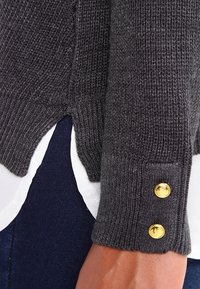 Tummanharmaa neulepusero, jossa on ribbikudontaa ja sivuhalkio. Kultaiset nappidetalit hihassa tuovat yksityiskohtia suunnitteluun.