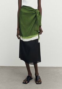 Lange groene wikkelrok met een zwart-wit patroon aan de onderkant, met een geknoopte taille. Gecombineerd met zwarte slidesandalen.