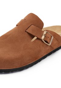 Chaussure slip-on en daim marron au design minimaliste, dotée d'une boucle en métal et d'une semelle en caoutchouc noir texturé pour l'adhérence.