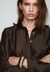 Camisa marrón de botones con un patrón de tachuelas plateadas y una textura suave. La modelo lleva una pulsera de oro texturizada en su brazo.