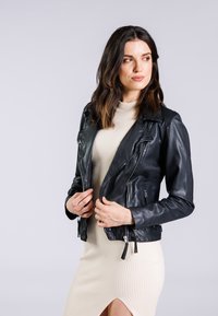 Schwarze Leder-Bikerjacke mit Reißverschlusstaschen, eingeschnittenem Kragen und strukturierter Oberfläche, getragen über einem hellen gerippten Kleid.