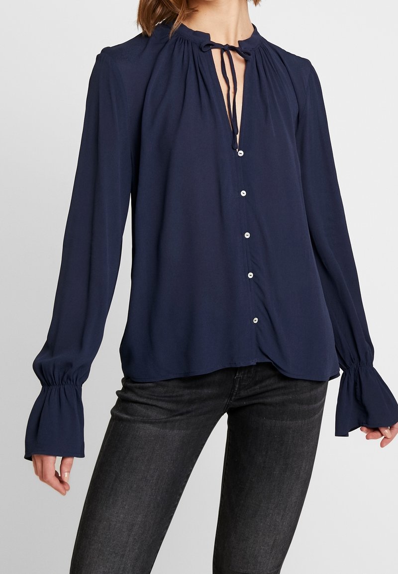 Navyblauwe blouse met lange mouwen, hoge hals, strikdetail, knoopsluiting en uitlopende mouwen. Glad materiaal met een lichte glans.