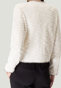 Biała kurtka o fakturowanej powierzchni z tkanym wzorem i frędzlami przy dolnym wykończeniu oraz rękawach, w zestawieniu z czarnymi spodniami, widoczna z tyłu.