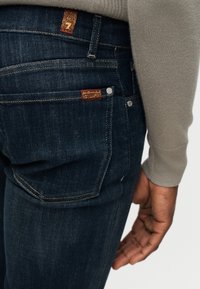 Κοντινό πλάνο μέσης ατόμου που φοράει σκούρο μπλε τζιν με κόκκινη και κίτρινη ετικέτα μάρκας και μπεζ πουλόβερ με ριγέ μανίκι.