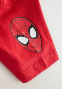 Tricou roșu din bumbac, cu un imprimeu al măștii Spider-Man alb-negru. Materialul este neted, cu o textură ușor granulată și guler rotund.