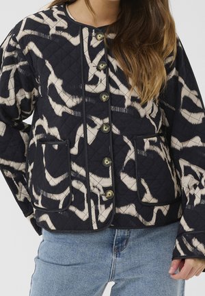 Quiltet jakke med sort og beige abstrakt mønster, rund halsudskæring, store knapper, to forlommer, båret med blå jeans.