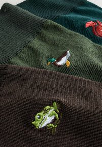 Trīs adītas cepures zaļos un brūnos toņos, ar izšūtiem zoss, vēža un vardes dizainiem. Materiāli ir strukturēti un riboti.