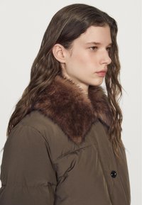 Giovane donna con lunghi capelli castani che indossa un giubbotto imbottito verde oliva con un ampio colletto di pelliccia marrone, rivolta a destra su uno sfondo neutro.