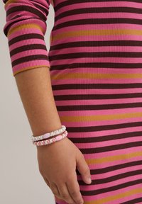 Randig ribbad klänning i rosa, brunt och gult; handen prydd med tre flätade armband i pastellfärger och pärlor.