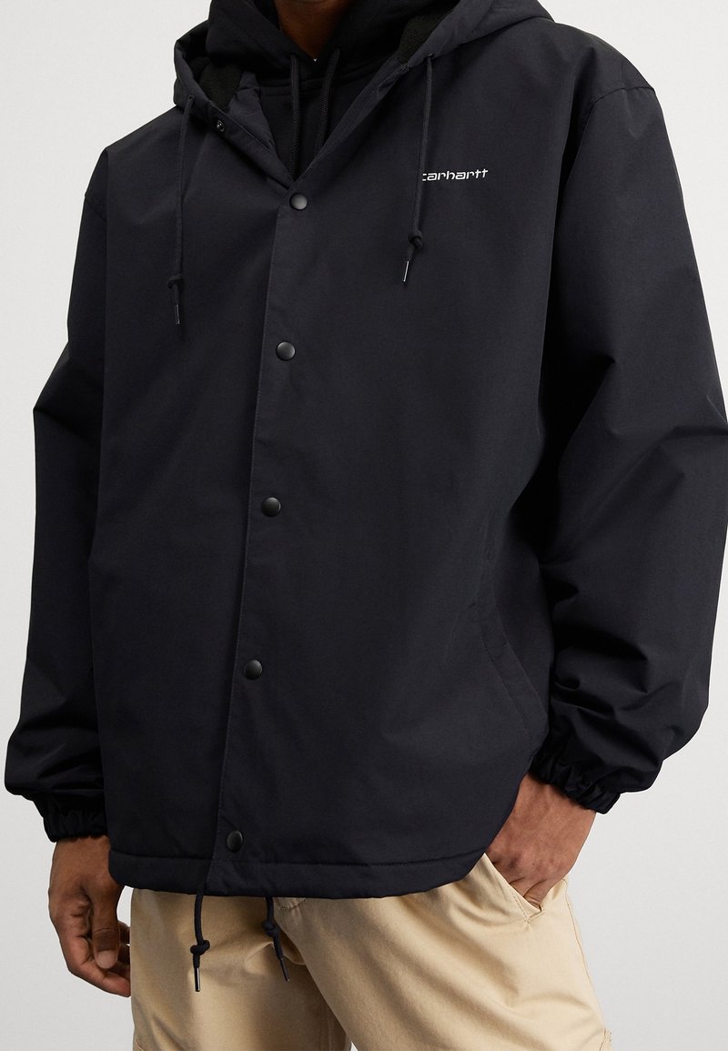 Man i en svart Carhartt jacka med huva och tryckknappar, samt beigefärgade byxor, med händerna delvis i fickorna, mot en neutral bakgrund.