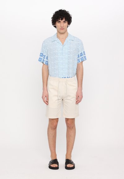 Cămașă cu mânecă scurtă de culoare albastru deschis, cu un model floral, asortată cu pantaloni scurți bej și șlapi negri. Materialul pare ușor și casual.