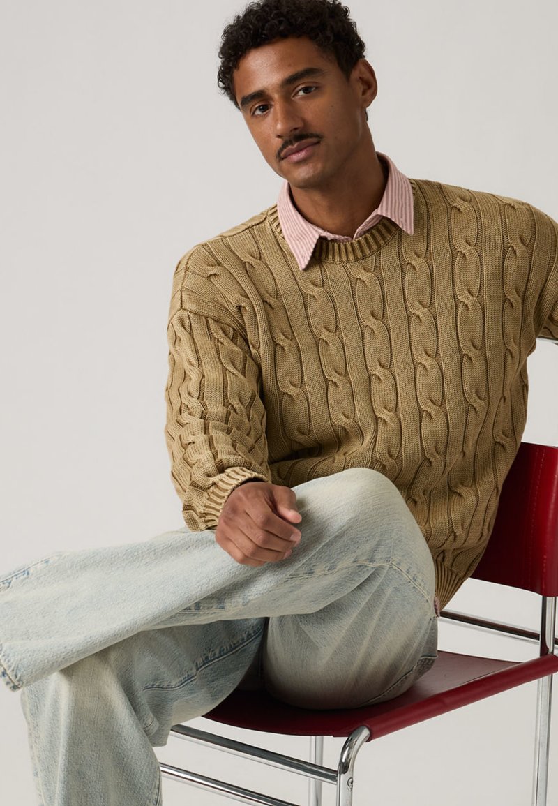 Man with short curly hair and mustache wearing beige cable-knit sweater over pink shirt, light-wash jeans, sitting cross-legged on red chair.