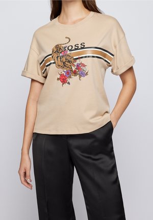 Chemise beige à manches courtes avec poignets retroussés, avec un motif tigre et fleurs colorées au-dessus de rayures noires, blanches et marron, portée avec un pantalon noir.