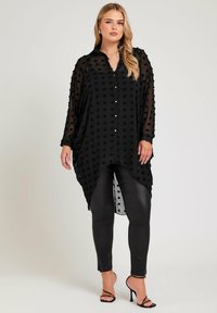 Svart genomskinlig skjorta med texturerade prickar, knappfront, asymmetrisk fåll, kombinerad med svarta glänsande leggings och remsandaler.