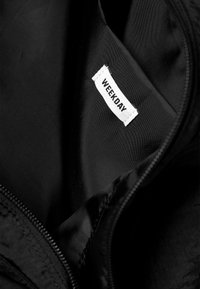 Black fabric interior with a visible white label reading "WEEKDAY." The texture appears smooth and slightly shiny, with a zipper detail.
