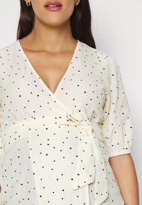 Blusa bianca con pois neri, design avvolgente, maniche corte a palloncino e vita legata in evidenza. Tessuto leggero e testurizzato.