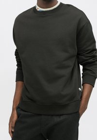 Homme portant un sweat-shirt vert foncé à col rond avec des poignets et une taille côtelés, associé à un pantalon foncé, main dans la poche.