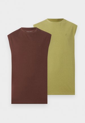 Homme debout les mains dans les poches, portant une chemise marron sans manches, un pantalon blanc et des chaussures marron à enfiler sur un fond blanc uni.